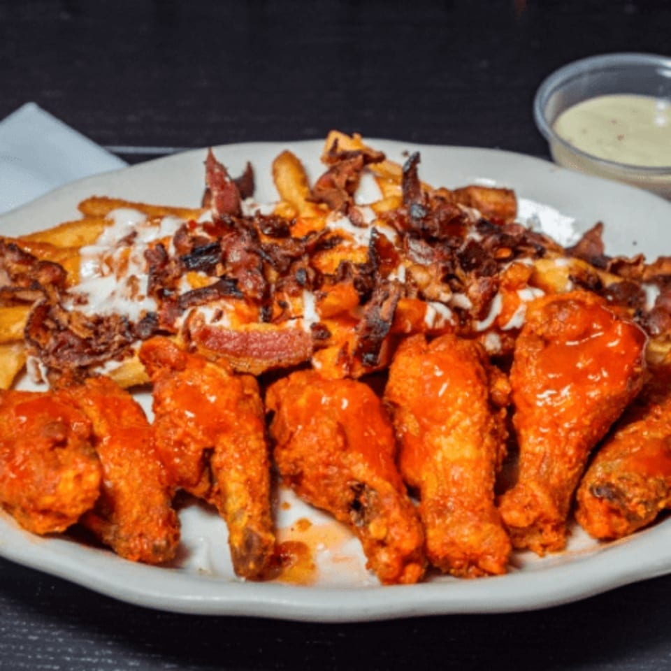 A plate of buffalo wings and loaded fries with bacon, cheese, and sauce, with a side of dipping sauce.