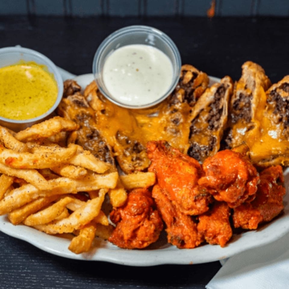 A platter of Jordan's Hot Dogs & Mac's fries, buffalo wings, and cheesesteak egg rolls with dipping sauces.