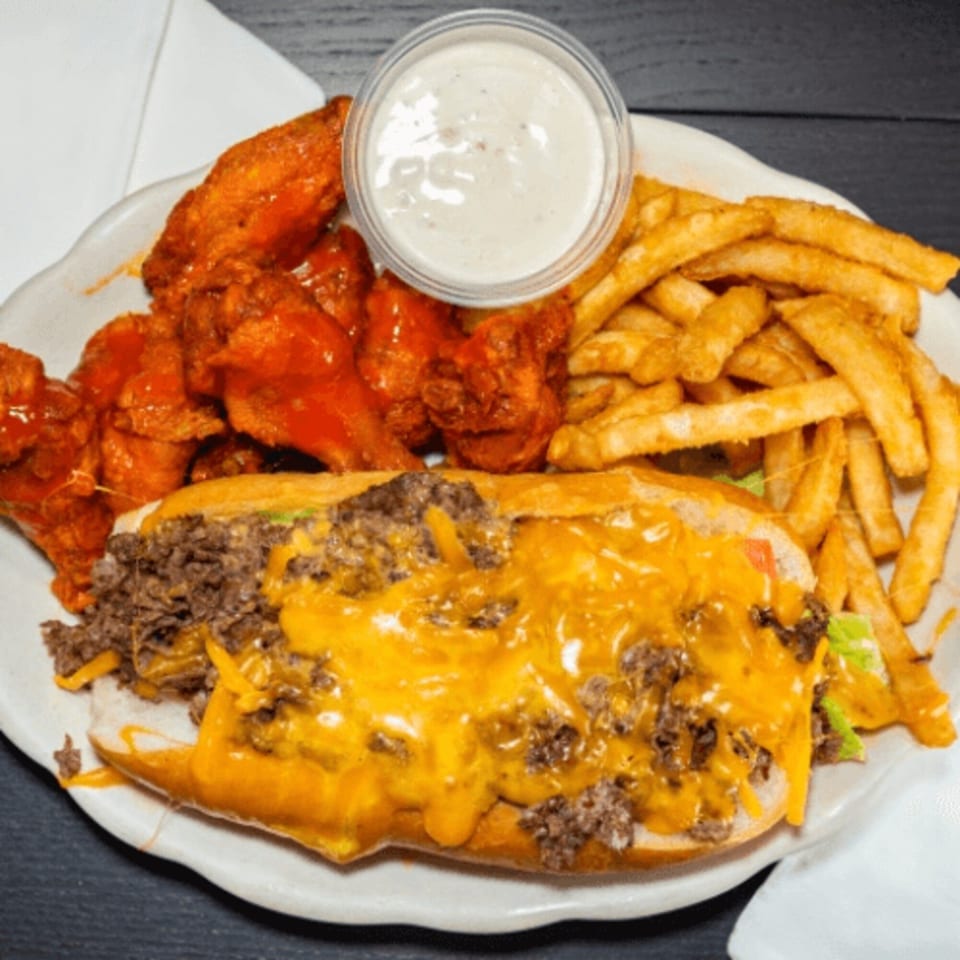 Cheesesteak sub with fries, buffalo wings, and a side of ranch dressing on a white plate.
